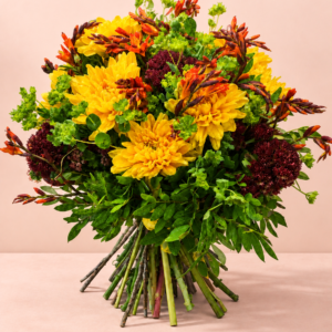 Ramo Alegría Redonda con flores amarillas, burdeos y naranjas sobre una mesa pastel con sombra natural